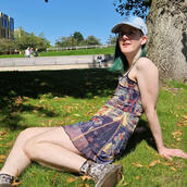 Mikey May, a white trans man with green hair wearing a blue cap and a sleeveless dress, is sat on grass in the sun.